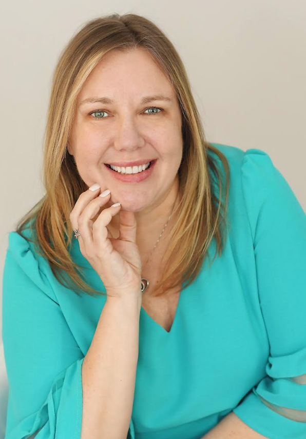 Portrait of a smiling woman with long dark curly hair, wearing a floral blouse and layered necklaces, against a light grey background.