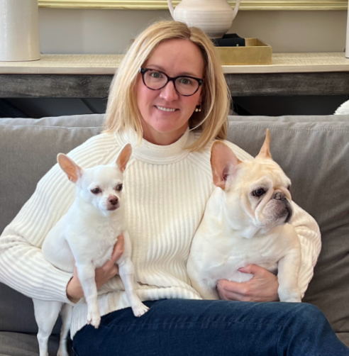 Portrait of a smiling woman with long dark curly hair, wearing a floral blouse and layered necklaces, against a light grey background.