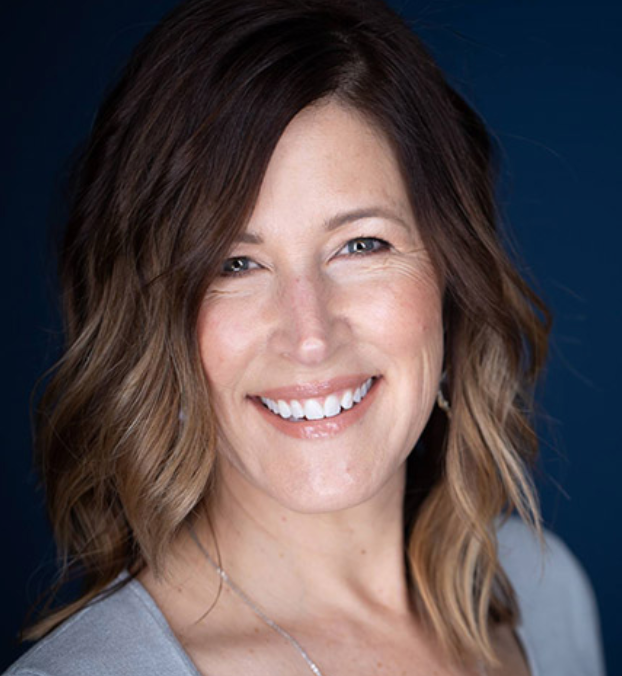 Portrait of a smiling woman with long dark curly hair, wearing a floral blouse and layered necklaces, against a light grey background.