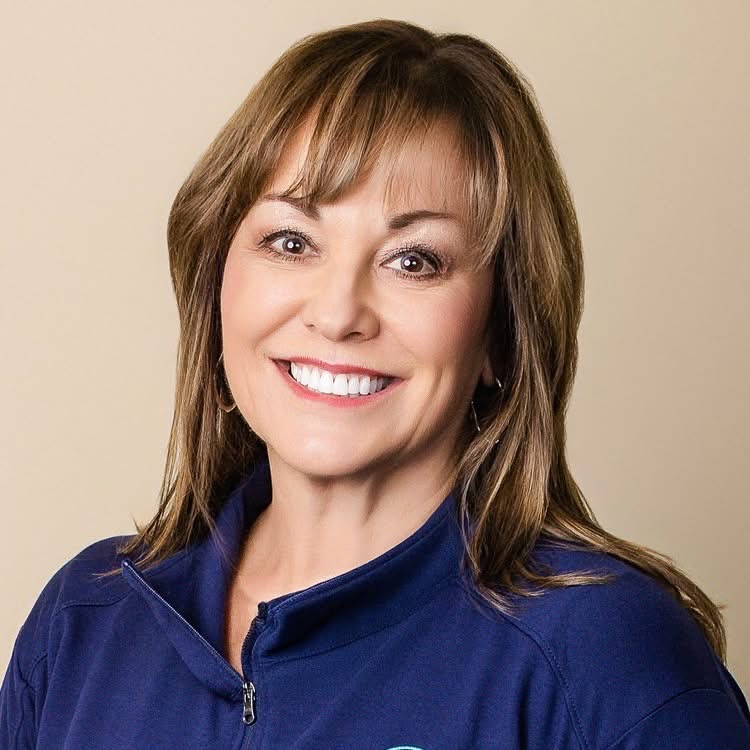 Portrait of a smiling woman with long dark curly hair, wearing a floral blouse and layered necklaces, against a light grey background.