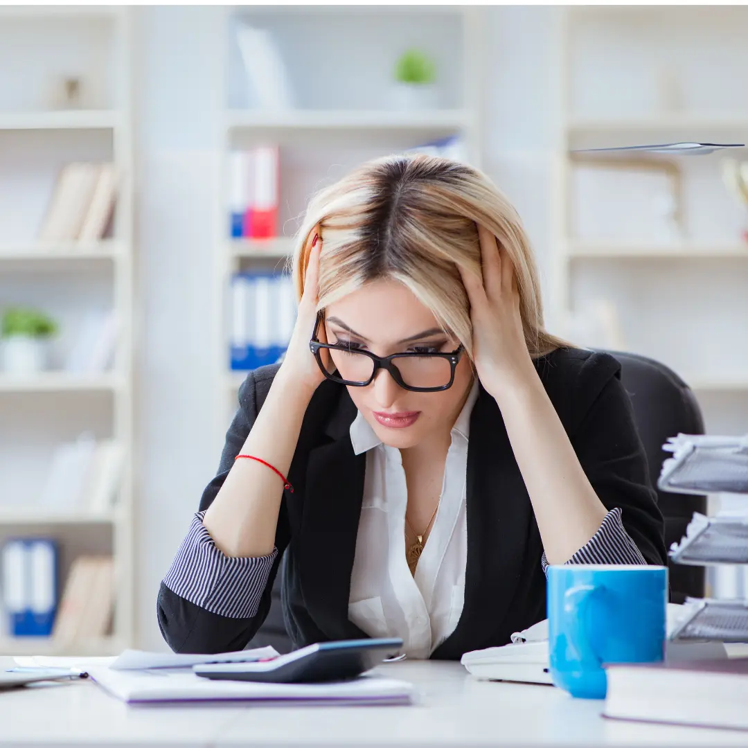 Stressed blonde woman in glasses at an office desk holding her head, surrounded by paperwork and a calculator