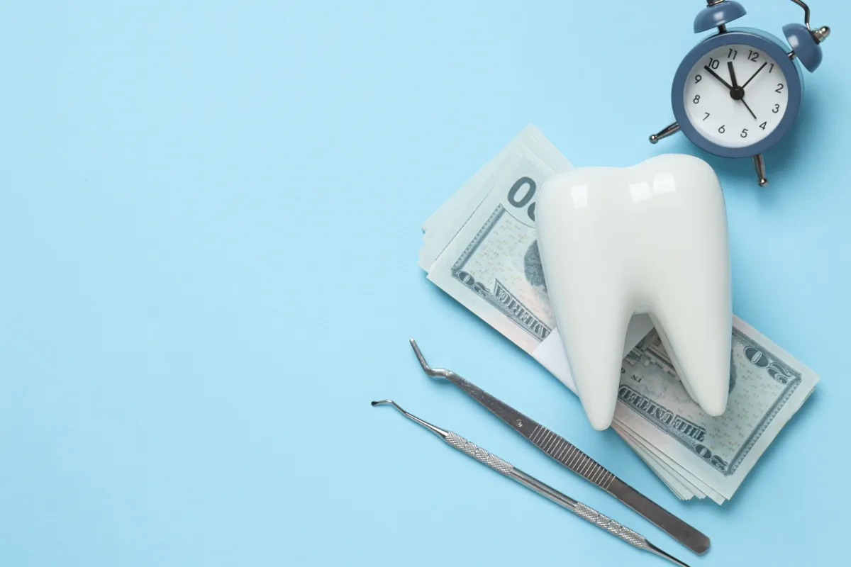Ceramic tooth model propped on a stack of US dollar bills beside dental instruments and a blue alarm clock on a light blue background