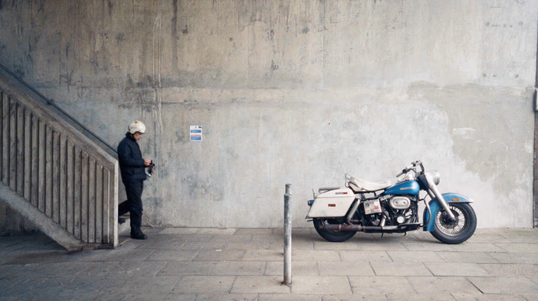 Person wearing a helmet and dark clothing standing near stairs next to a vintage blue and white motorcycle against a concrete wall.