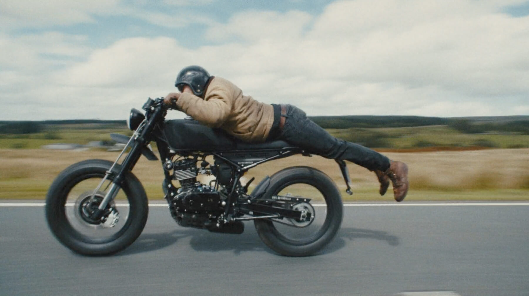 Person wearing a helmet and jacket lying flat on a black motorcycle while riding on a road with a blurred countryside background.