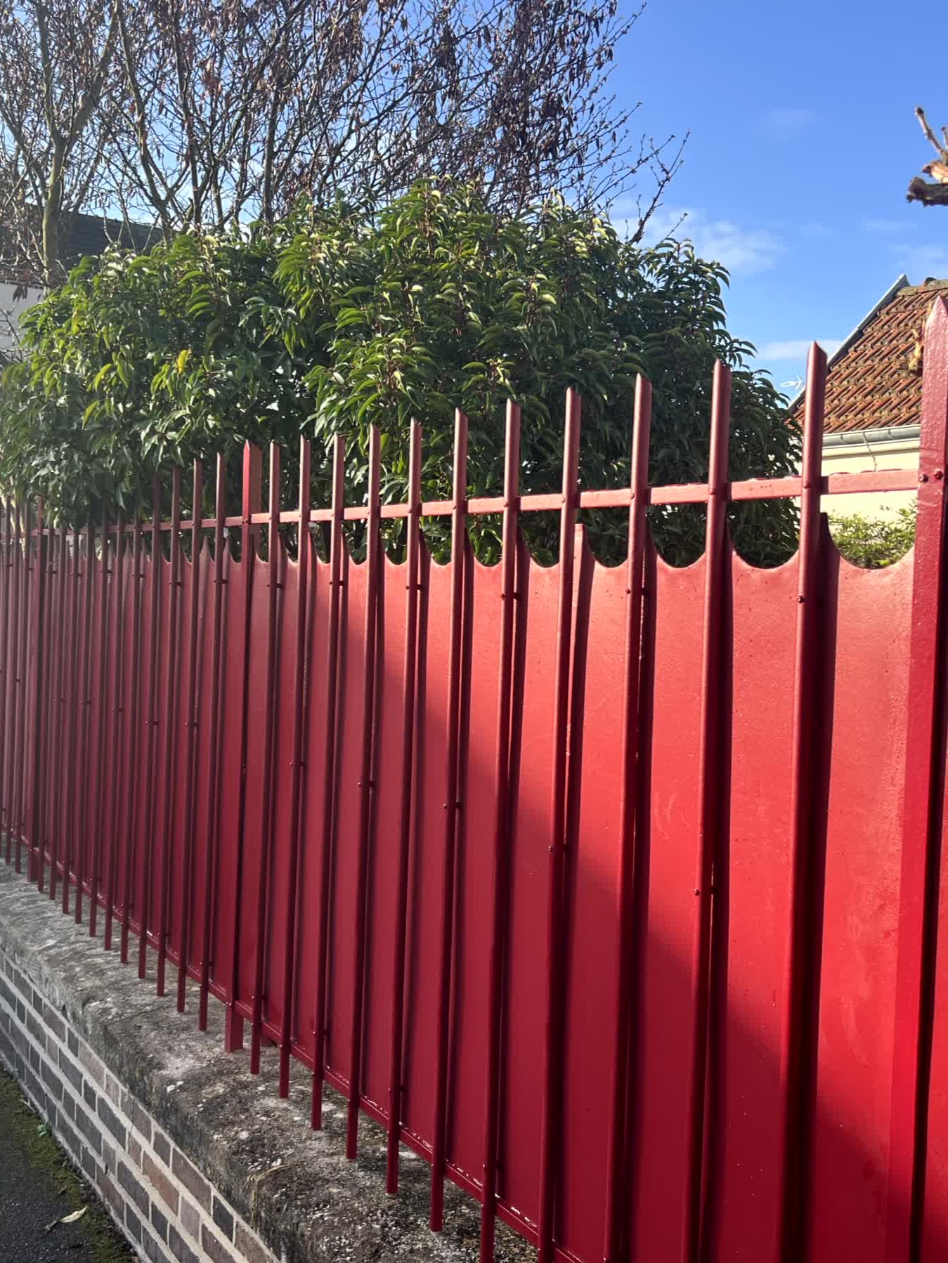 Clôture métallique rouge avec des pointes verticales devant un buisson vert sous un ciel bleu.
