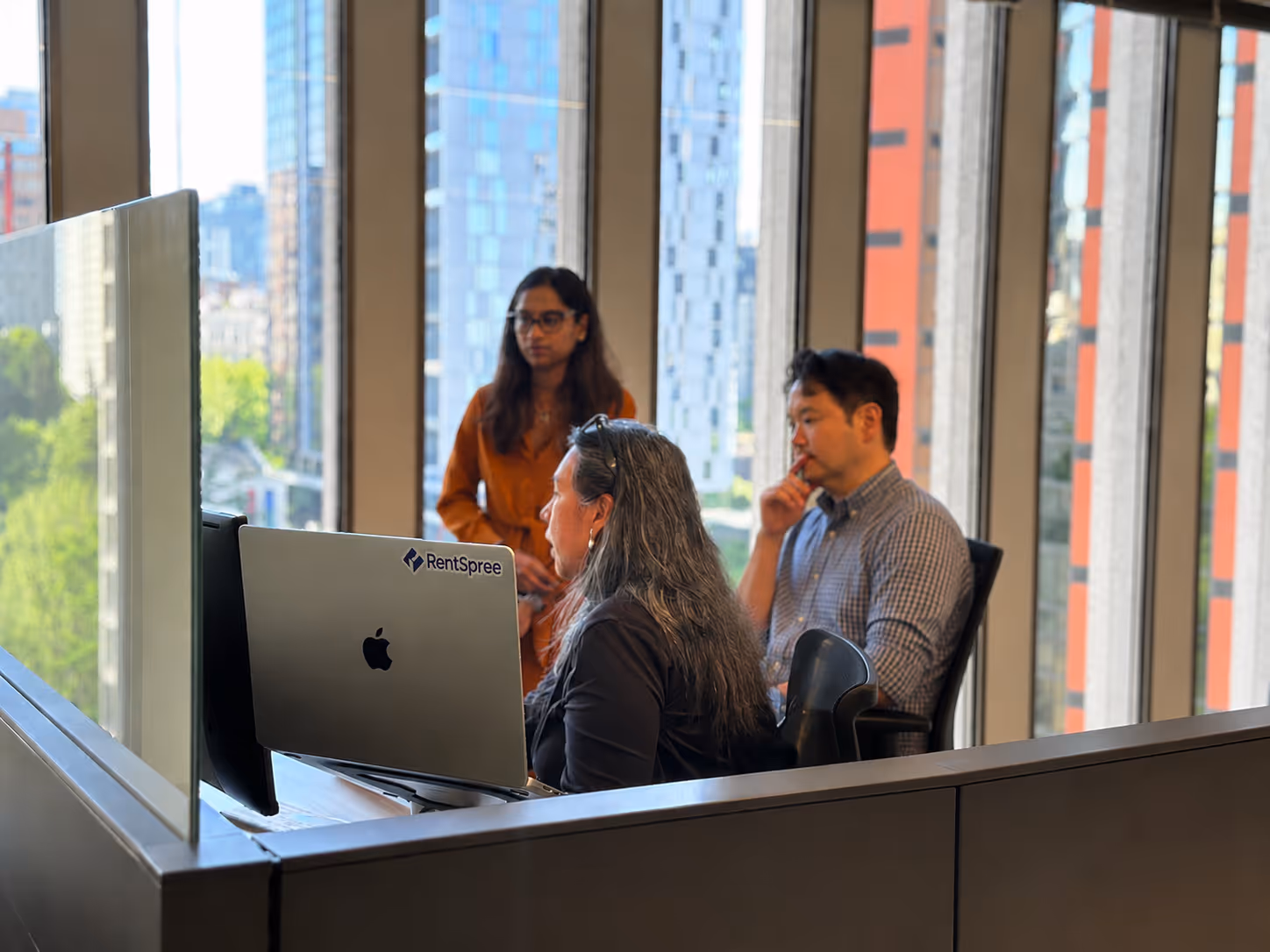 Three colleagues in an office with large windows, one seated at a laptop labeled RentSpree.