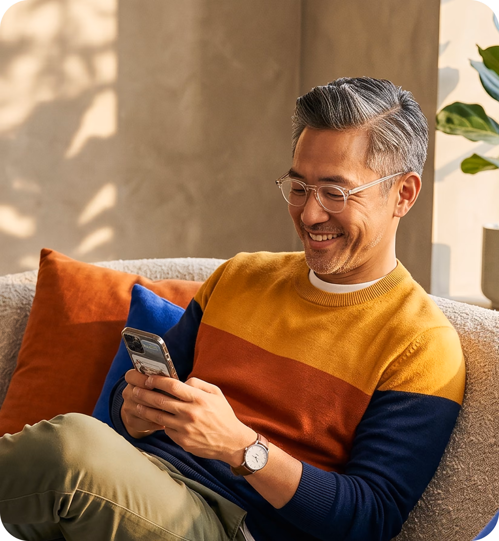 Smiling man with glasses in color-block sweater using smartphone on beige couch with cushions.