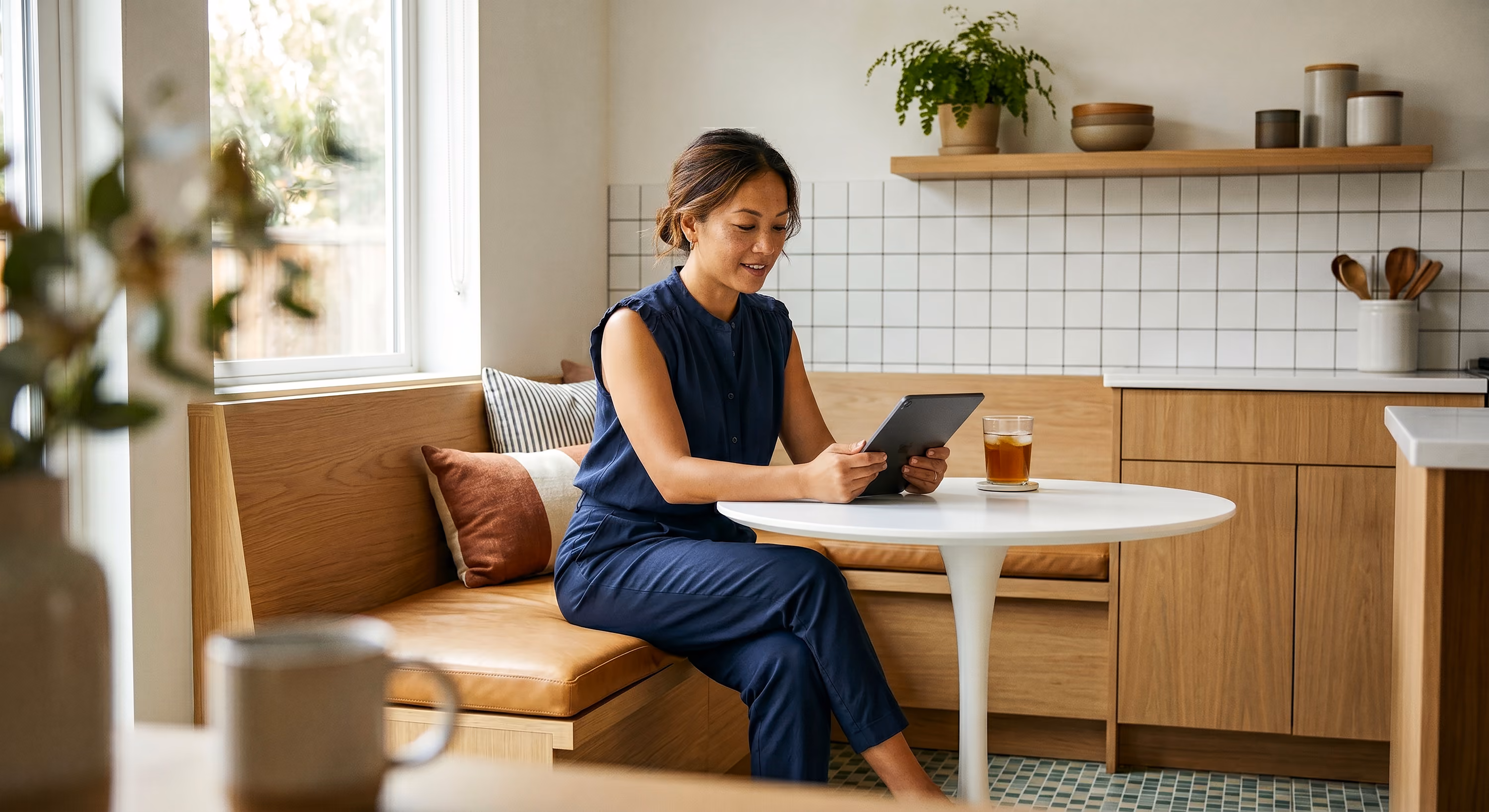 Woman in navy outfit sitting on wooden bench reading a tablet in a modern kitchen nook.