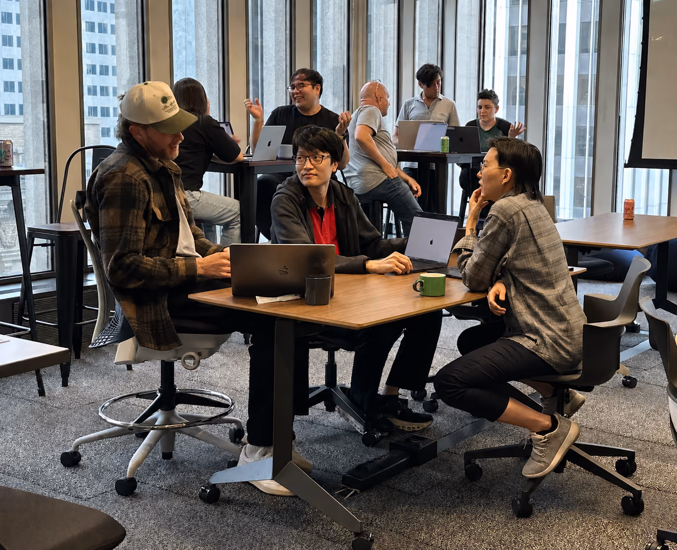 A diverse group of people working and chatting at laptops in a modern office with large windows.