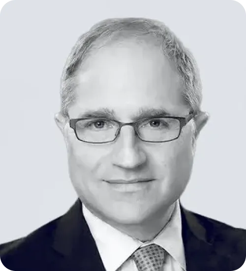 Portrait of a middle-aged man wearing glasses, a suit, white shirt, and patterned tie, smiling slightly against a plain light background.