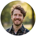 Smiling man with wavy brown hair and beard wearing a plaid shirt outdoors with blurred greenery background.