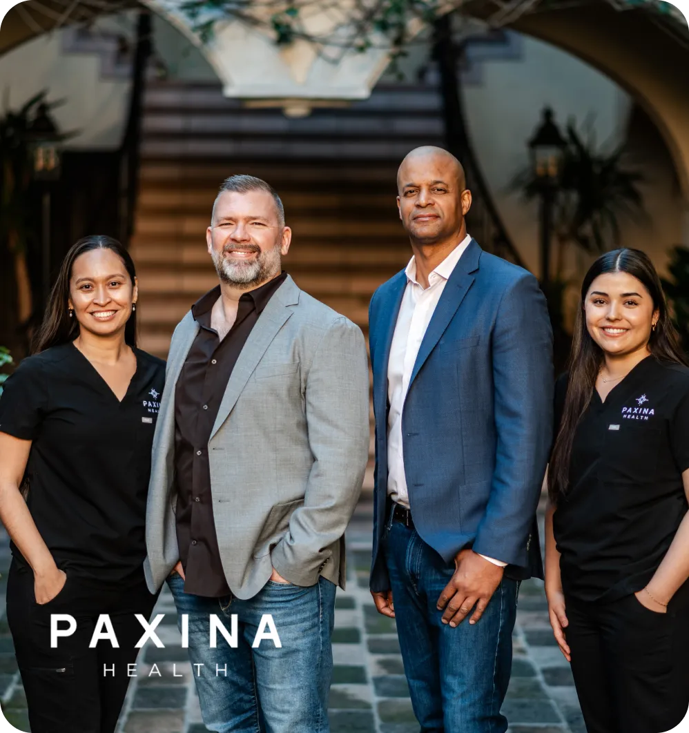 Four professionals standing in front of a staircase, two women in black Paxina Health scrubs and two men in business casual attire.