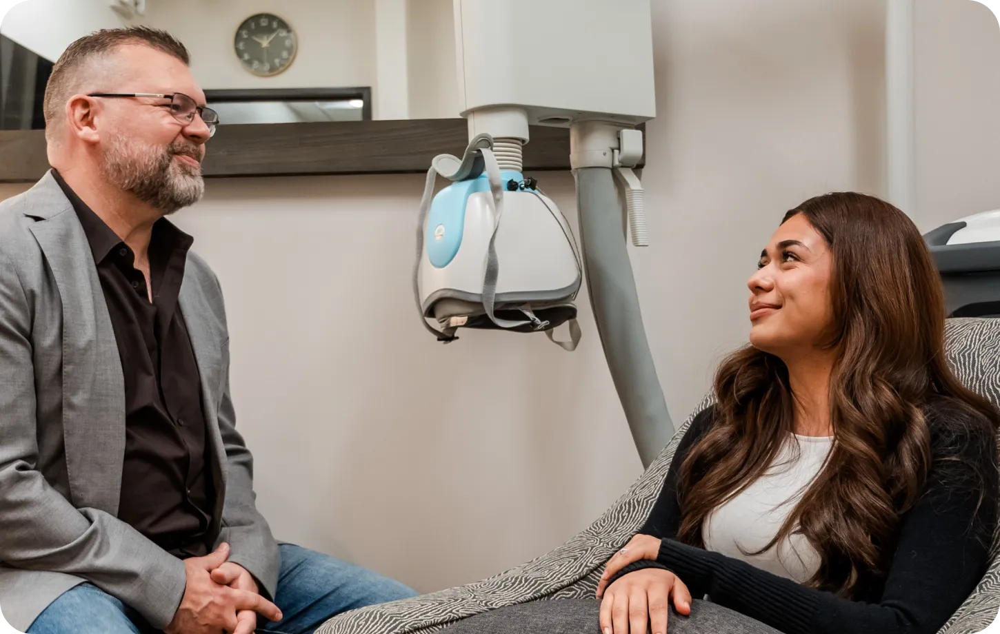 Man with glasses and beard in grey blazer sitting and smiling at a woman reclining on a chair in a clinical setting with medical equipment nearby.
