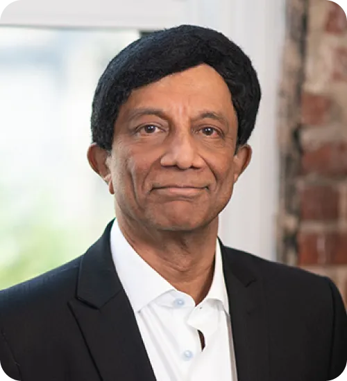 Middle-aged man with black hair wearing a black suit jacket and white shirt, smiling slightly against a window and brick wall background.