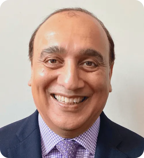 Smiling middle-aged man wearing a dark suit jacket and a purple patterned shirt with a matching tie against a plain light background.