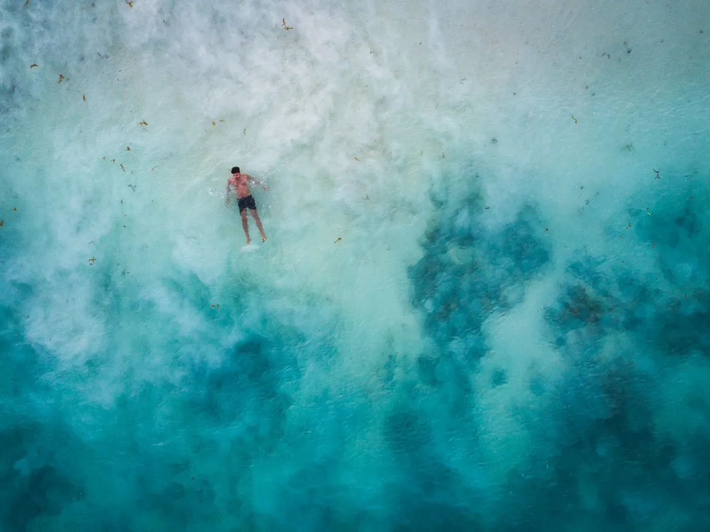 A man in board shorts floats in the middle of the shallow, clear waters of the ocean.