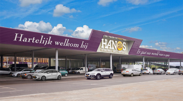 Exterior view of Hanos wholesale store with purple signage on the roof saying 'Hartelijk welkom bij HANOS' and 'Er gaat een wereld voor u open' under a partly cloudy blue sky, with a parking lot of several cars in front.