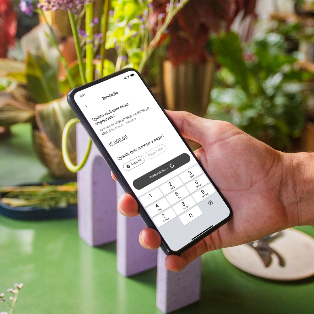 Pessoa segurando um smartphone em um ambiente com muitas plantas. O smartphone exibe uma tela de aplicativo financeiro mostrando um valor de R$ 4.200,00 e opções relacionadas a pagamentos. A pessoa parece estar revisando ou confirmando a requisição de um empréstimo sem burocracia.