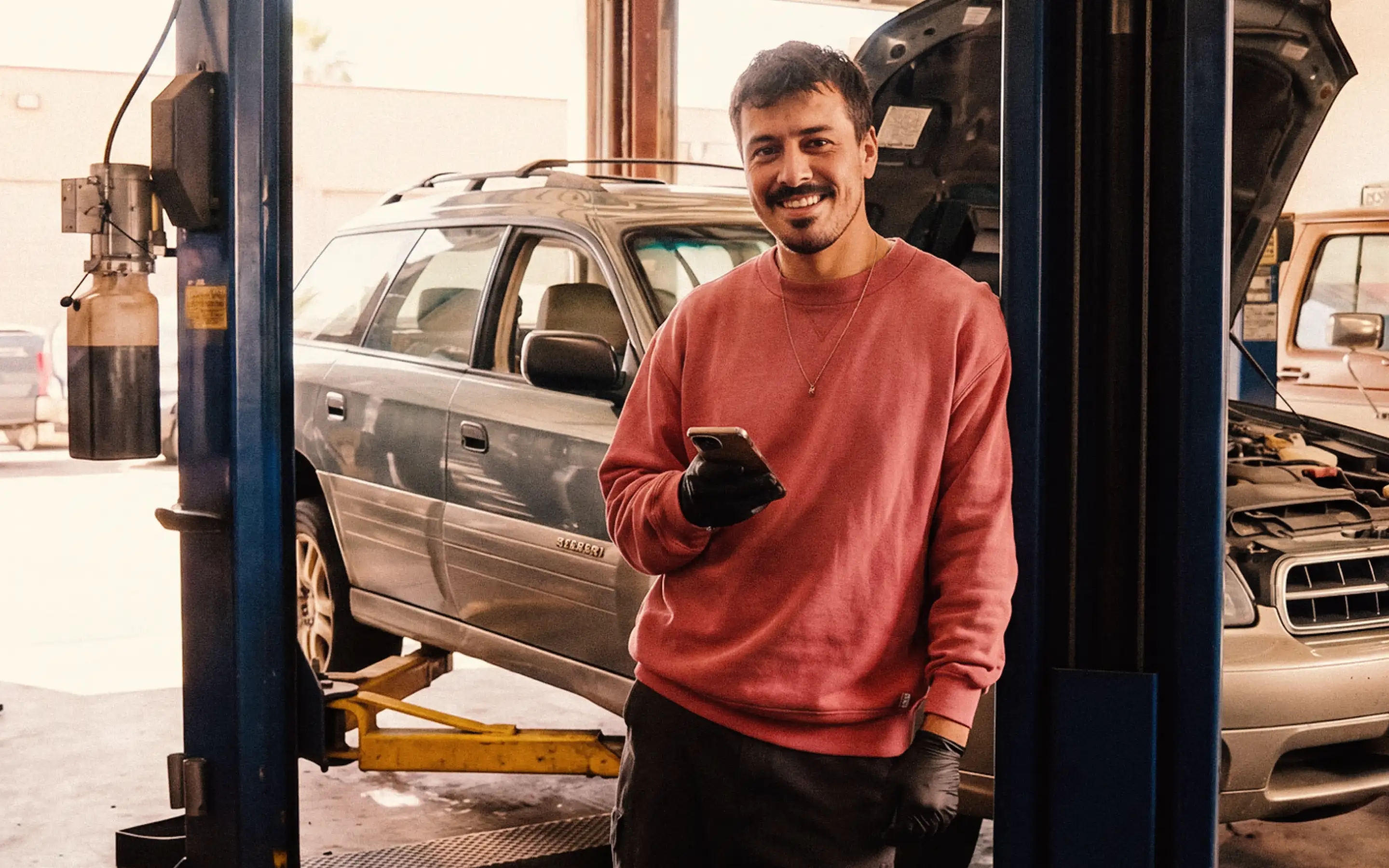 Man in red sweatshirt and black gloves smiling, holding phone, standing in a garage by a car on a lift.