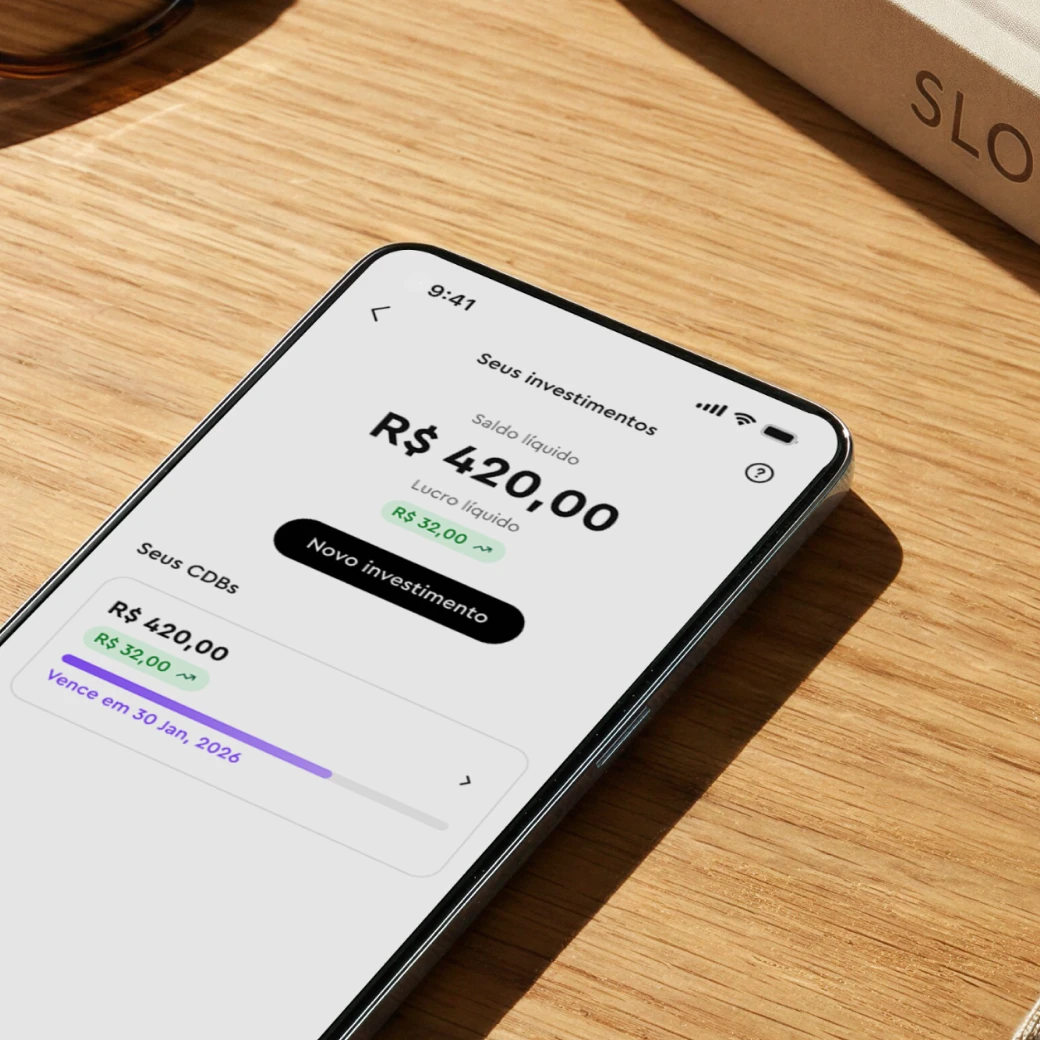 Smartphone on wood table showing investment app with R$420 balance and R$32 profit in CDBs.