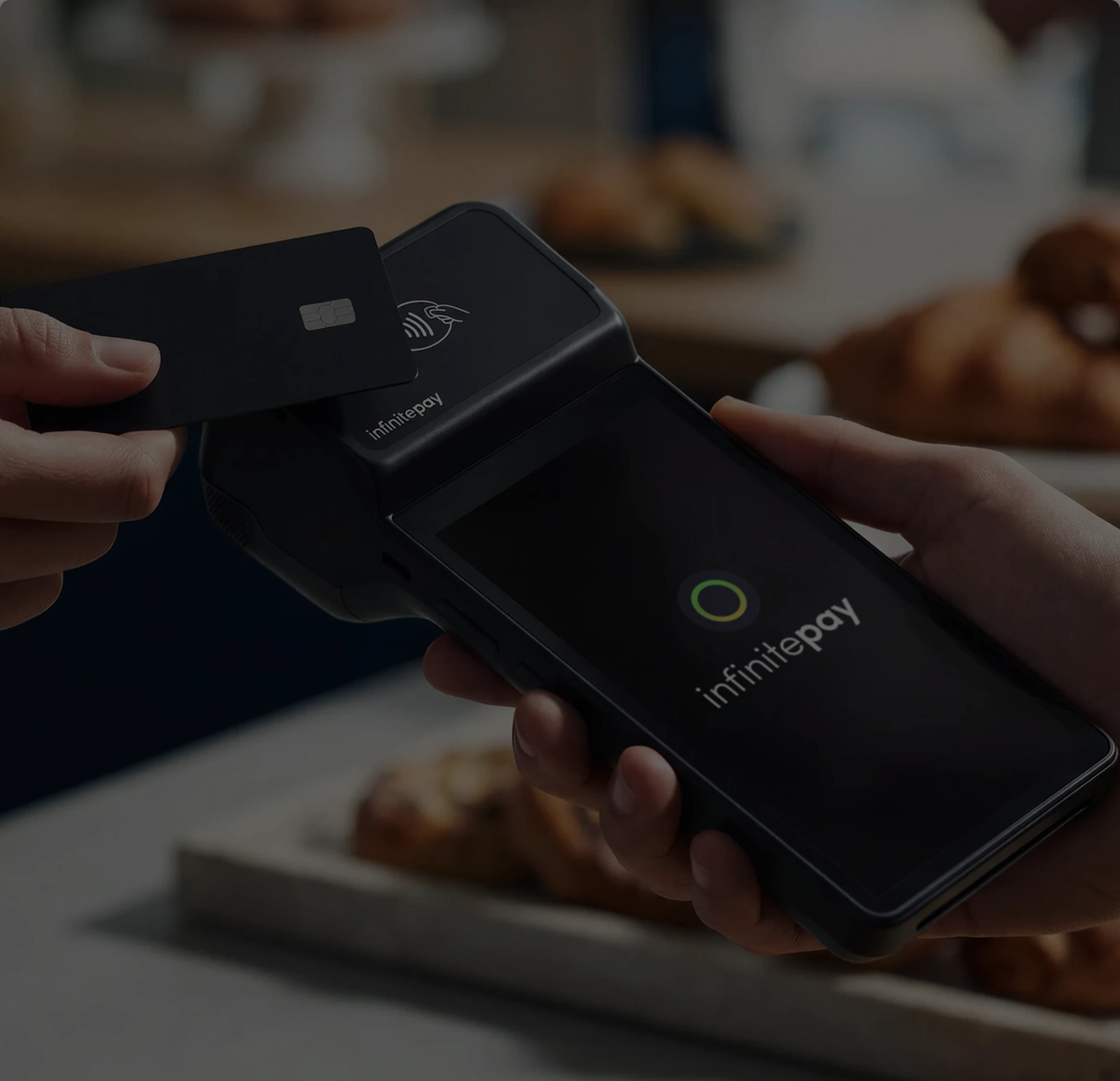Pessoa segurando leitor de cartão InfinitePay enquanto outra aproxima cartão preto para pagamento.