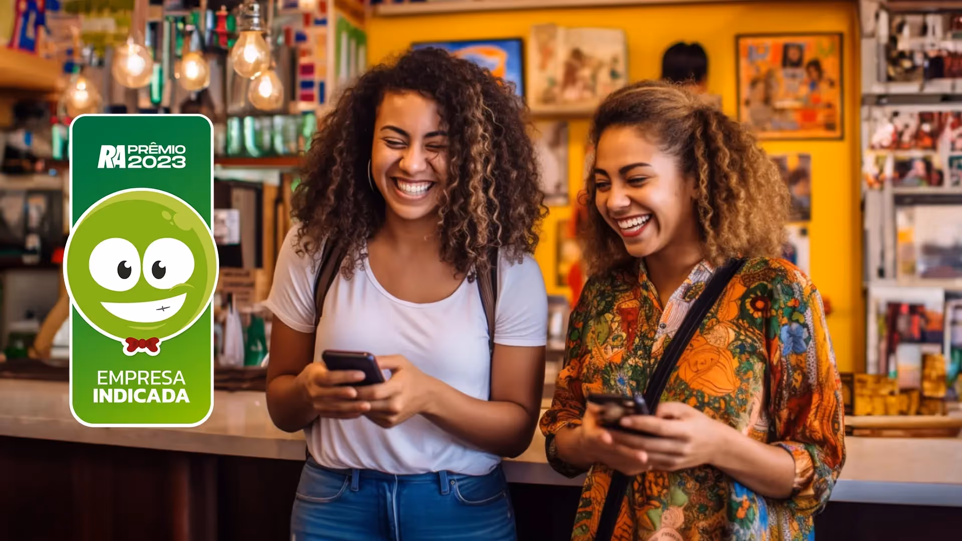Duas mulheres negras sorrindo com celular nas mãos. A mulher a esquerda está com cabelo crespo com luzes loiras e preso, sua camisa é longa e estampada em tons laranjas e verdes. A outra possui cabelos crespos soltos, ela está de camisa branca e calça jeans azul. No canto da foto, há o símbolo do RA100 conquista da InfinitePay, por ser a empresa de maquininha mais bem avaliada na sua área de maquininha de cartão 