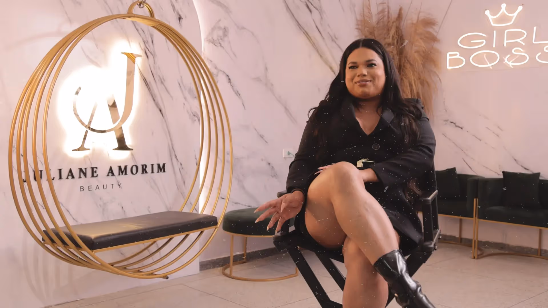 Imagem mostra a maquiadora Juliane Amorim em seu estúdio de maquiagem, sentada ao lado de um balanço dourado num fundo marmorizado