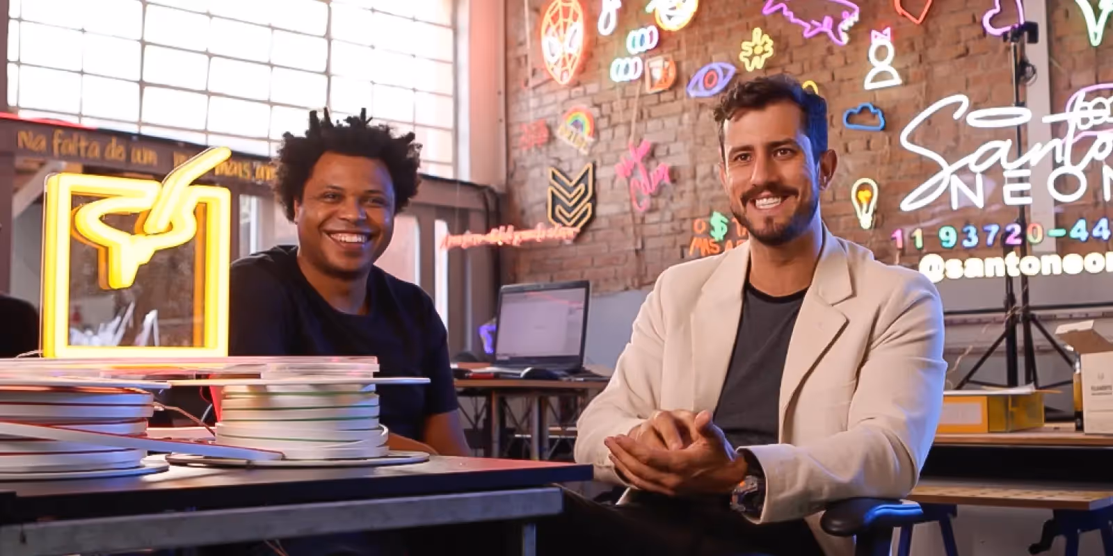 Dois homens sorridentes em um estúdio de neon, com várias luminárias e letreiros de neon coloridos ao fundo, incluindo o letreiro "Santo Neon".