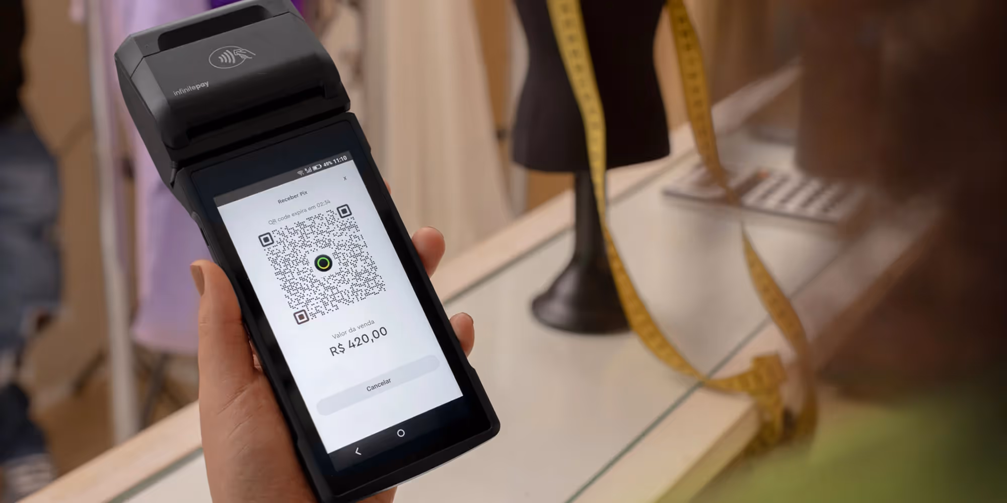 Foto que ilustra conteúdo sobre gerar QR Code Pix mostra uma maquininha de cartão com um QR Code no visor, com o valor de R$420,00 sendo cobrado. Uma mão feminina segura a maquininha, e ao fundo é possível ver manequins que indicam se tratar de um estúdio de costura
