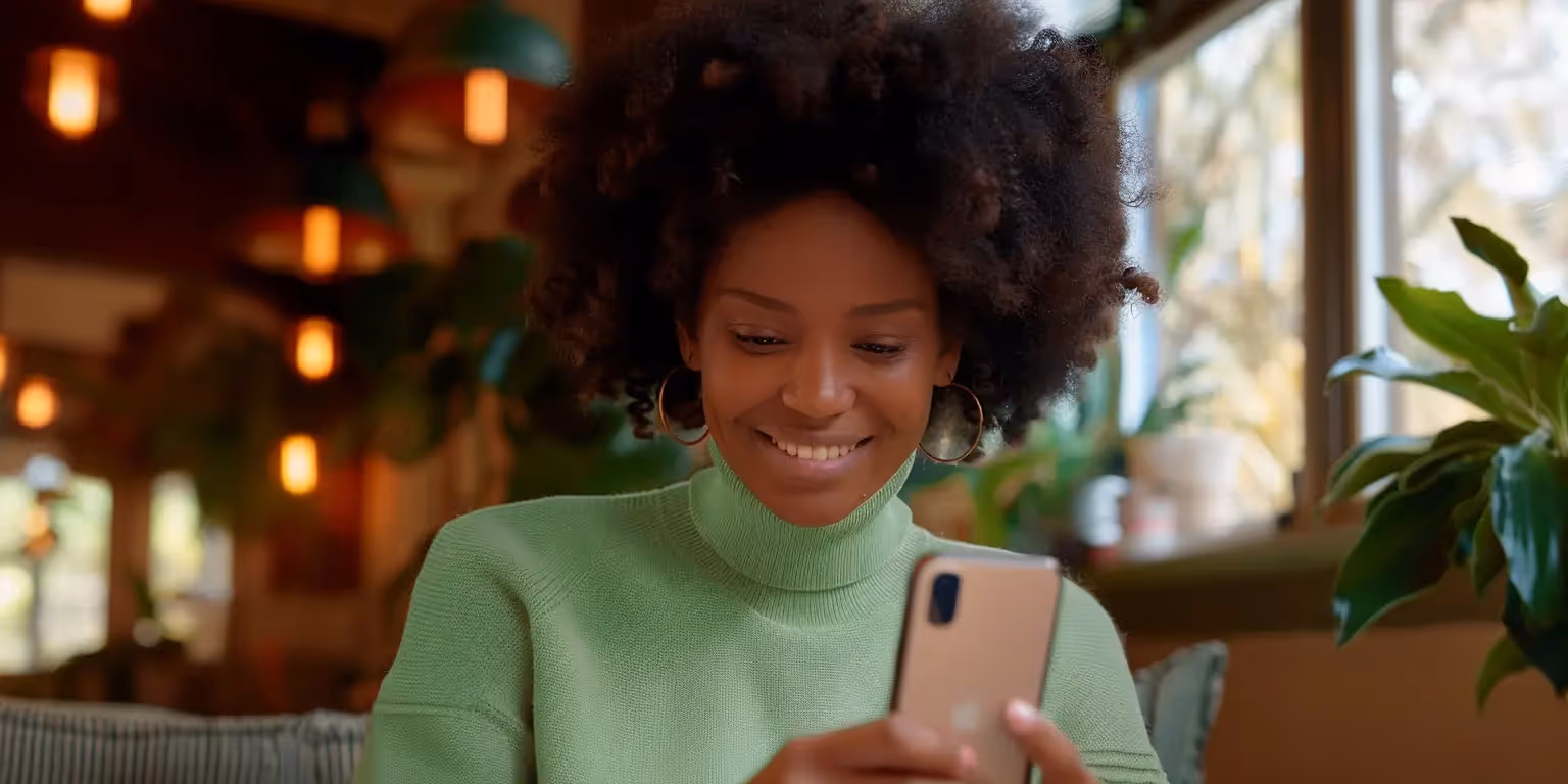 Mulher sorrindo enquanto olha para o celular, lendo sobre como funciona o Pix parcelado, vestindo um suéter verde em um ambiente acolhedor com plantas e luzes pendentes