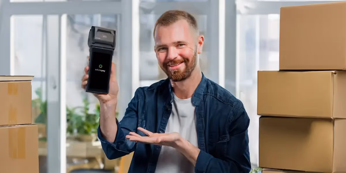 Homem está em seu escritório, rodeado de caixas de papelão, e mostra a maquininha InfinitePay para a câmera. Com a maquininha, ele consegue fazer inventário de estoque e otimizar a gestão dos seus produtos