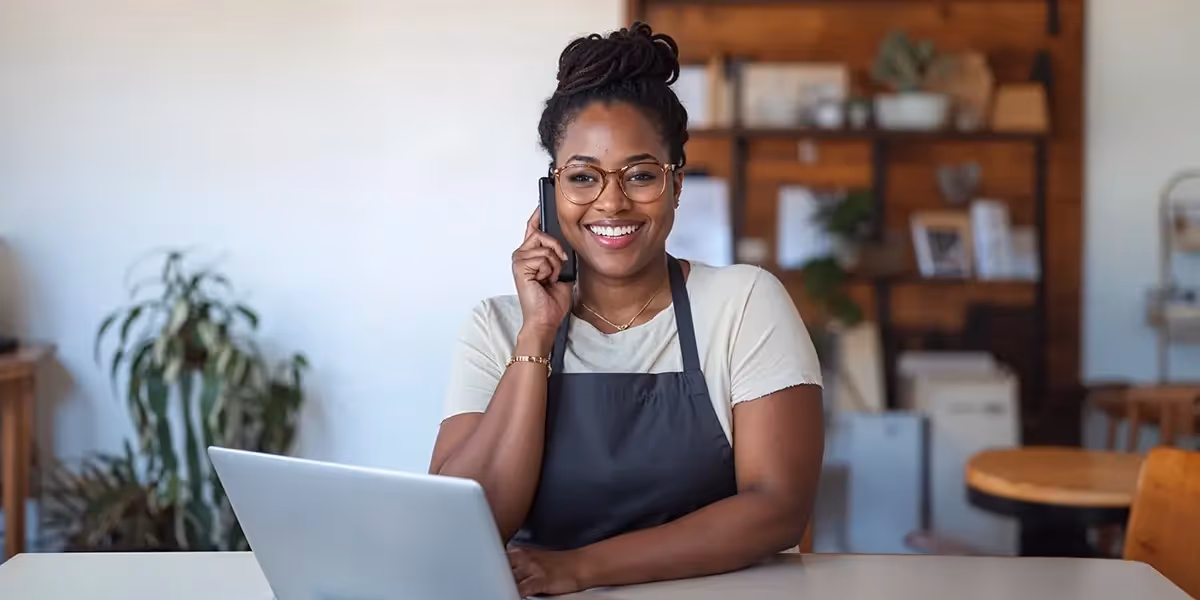 Mulher de avental sentada na frente de um computador com o telefone apoiado na orelha simbolizando "como cobrar um cliente"