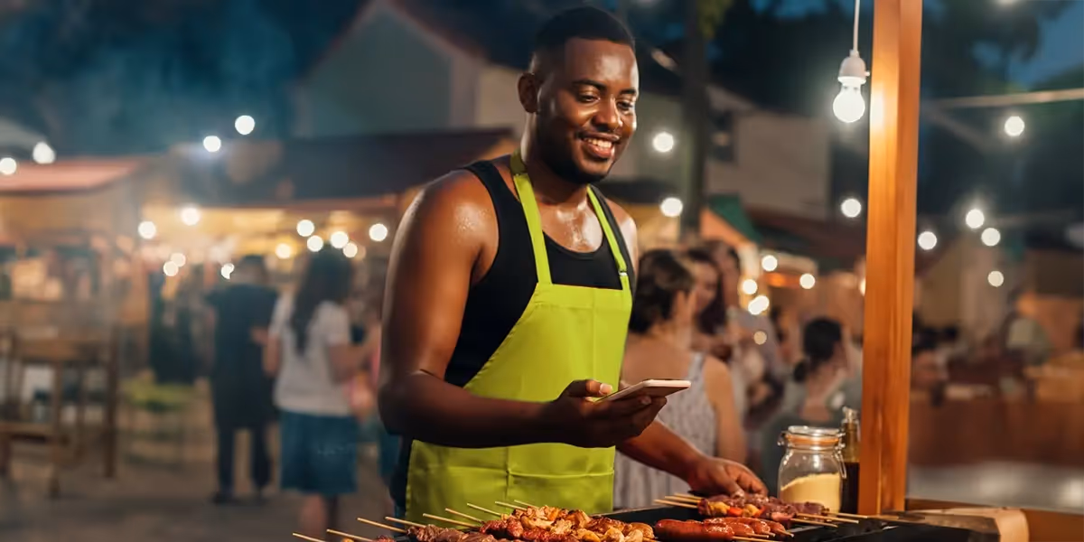 o que é renda informal: imagem mostra rapaz vendendo espetinhos em uma feira. ele está com o celular na mão, sorrindo.