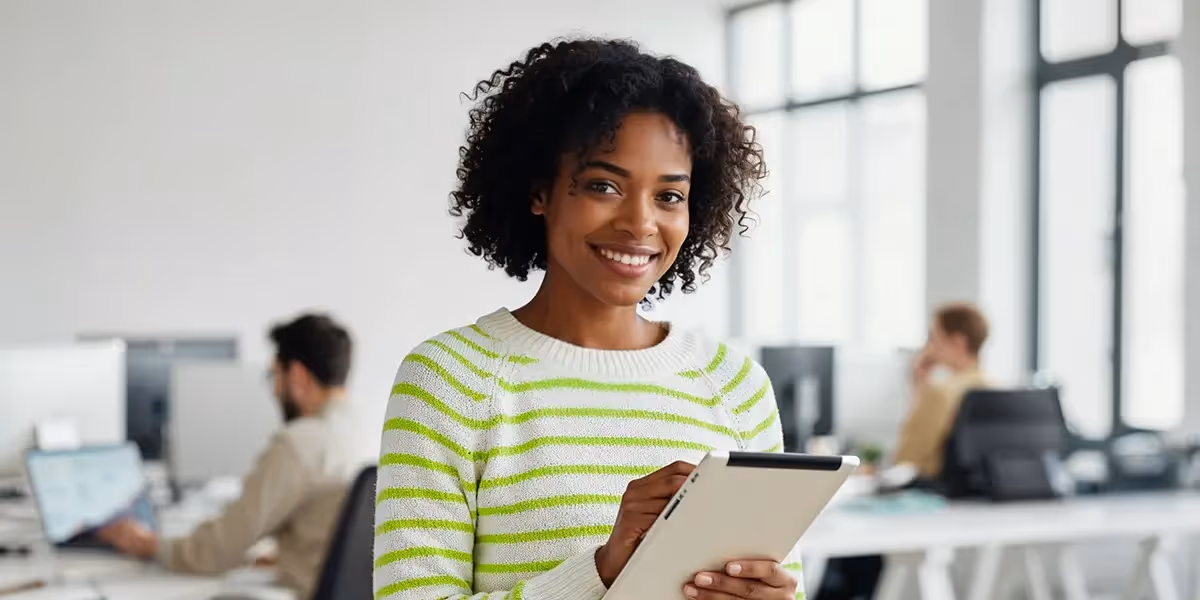 Mulher sorridente segurando um tablet em um escritório moderno, representando eficiência na gestão de canais de vendas. No fundo, colegas trabalham em computadores, reforçando um ambiente corporativo dinâmico
