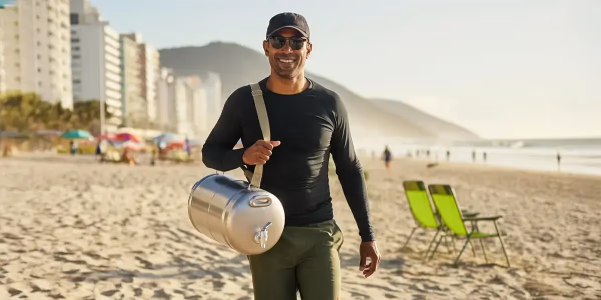o que vender na praia: homem sorridente carregando barril térmico de bebida na praia, sugerindo venda ambulante.
