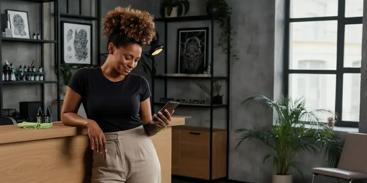 Mulher sorridente encostada no balcão de um estúdio, olhando o celular em ambiente moderno e iluminado.