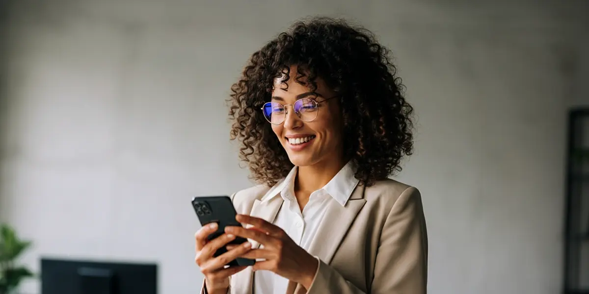 Mulher de óculos e cabelo cacheado sorrindo enquanto usa um smartphone para enviar whatsapp mensagem para clientes sumidos, vestindo um blazer bege sobre uma camisa branca.