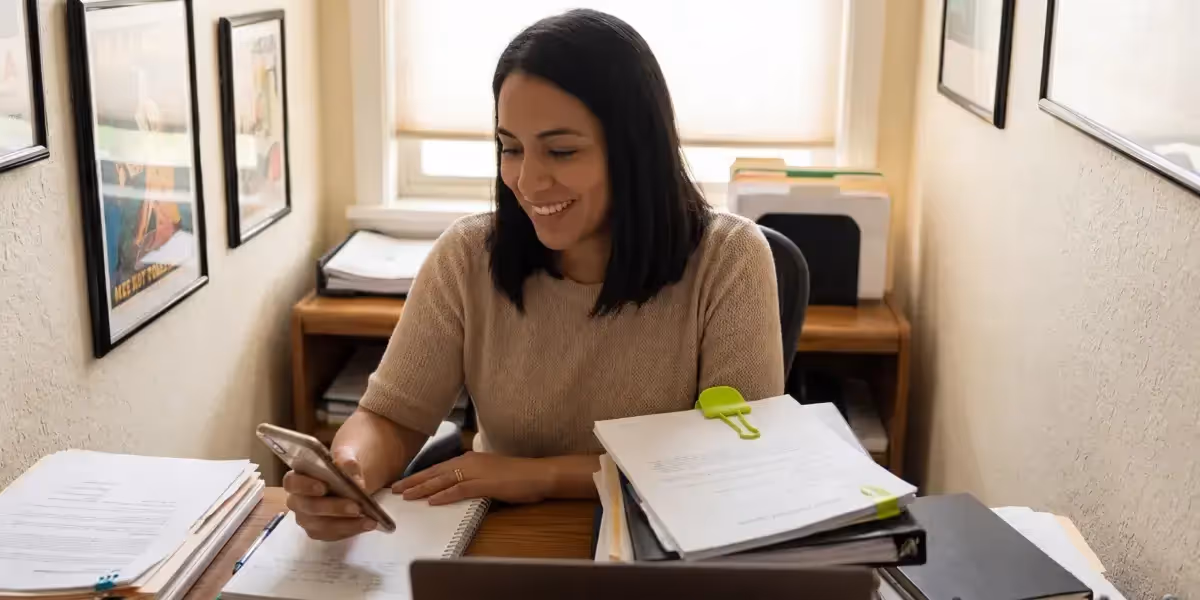 Mulher sorridente analisando no celular quanto rende 100 mil no CDB por mês em seu escritório.