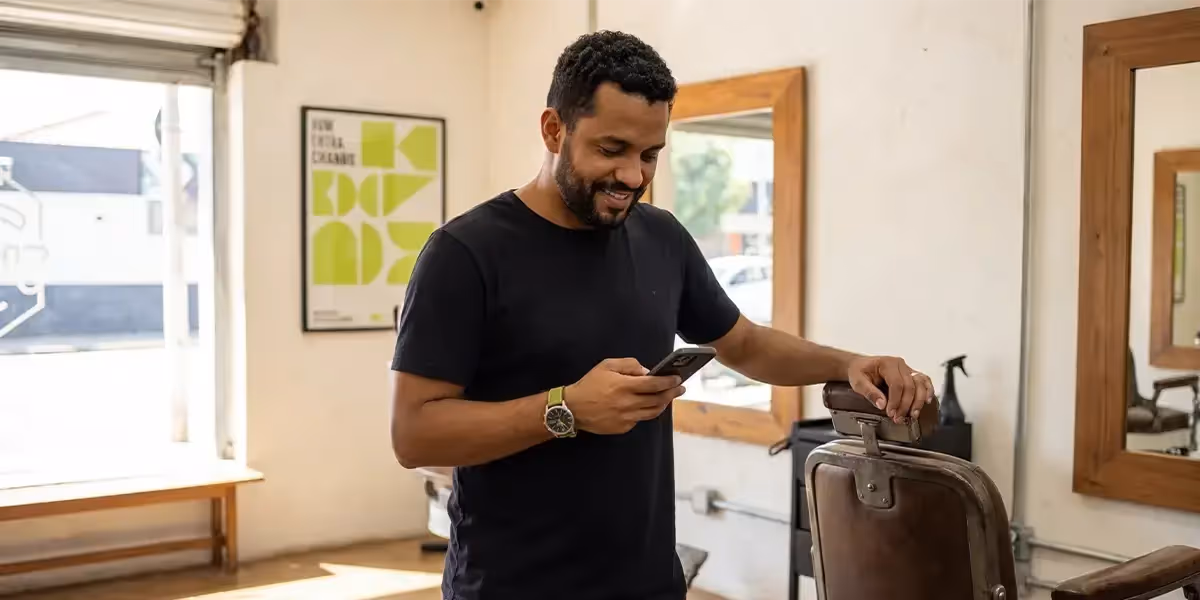 Homem sorrindo enquanto pesquisa no celular sobre prazo mínimo CDB em uma barbearia.