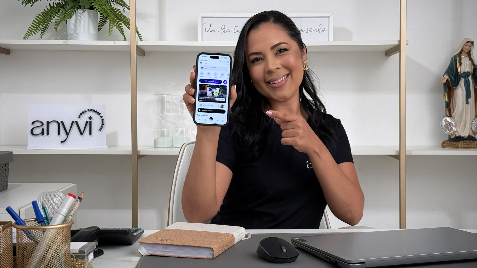 Woman sitting at desk smiling, holding and pointing at smartphone displaying an app screen.