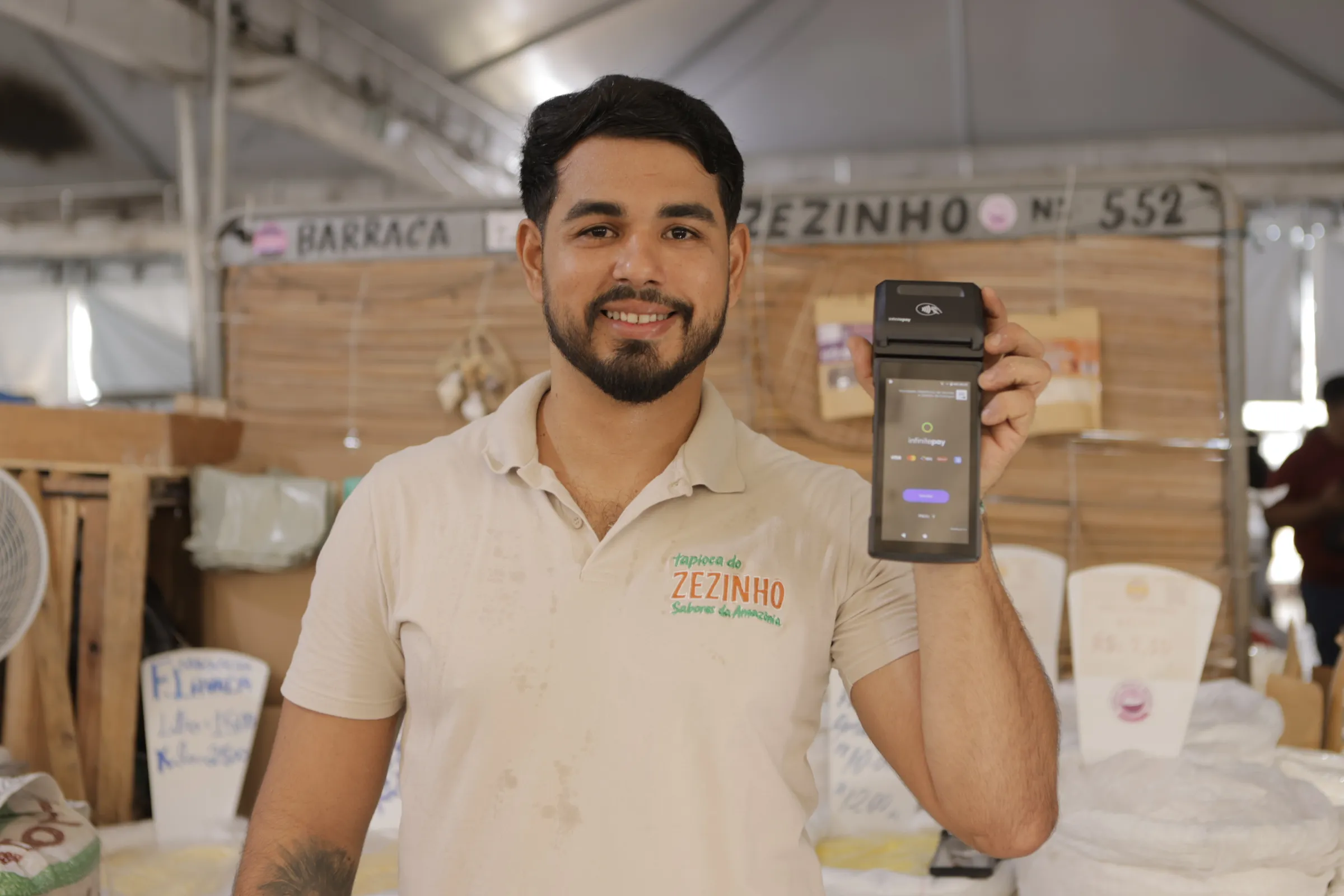 Smiling man in a beige shirt holding up a payment terminal in a market stall.