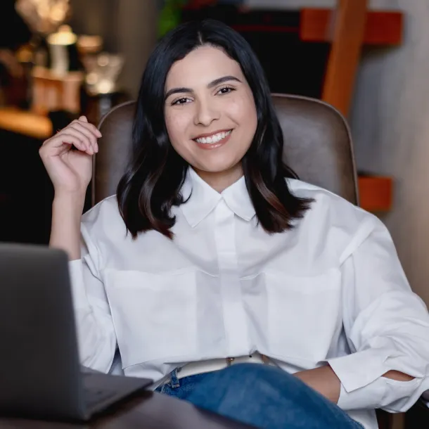Smiling woman with black hair wearing a white blouse sits casually in a chair by a laptop.