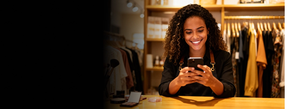 Smiling woman with curly hair using a smartphone at a wooden counter in a clothing store.