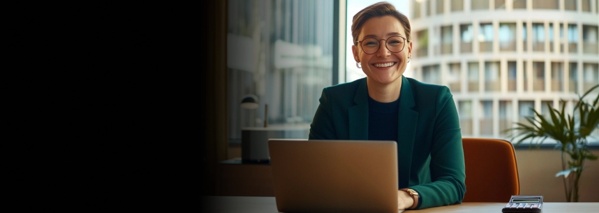 Smiling person with glasses working on a laptop at a desk in a modern office setting.