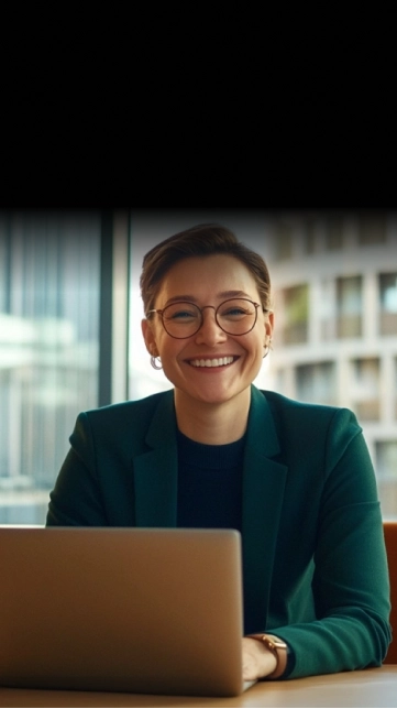 Smiling professional woman with glasses and short hair sitting at desk with a laptop in an office.