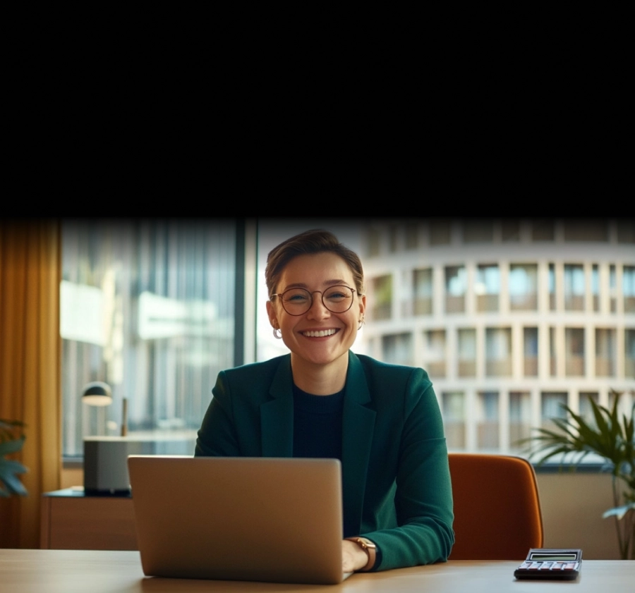 Smiling person with glasses sitting at desk with laptop and calculator in bright office.