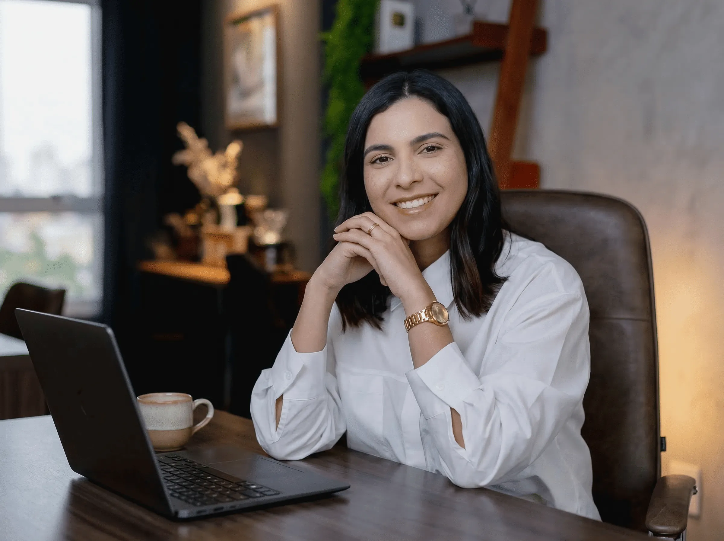 Smiling woman in white shirt sitting at desk with laptop and coffee cup in cozy home office.