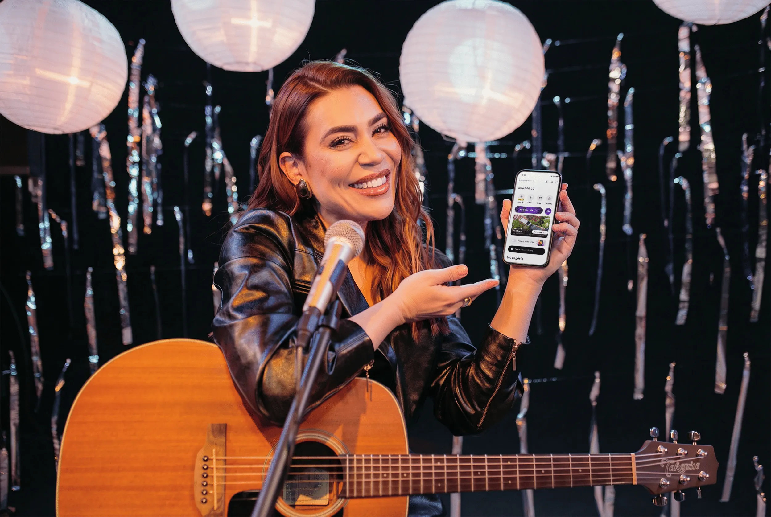 Smiling woman with guitar and microphone points to a phone screen showing a financial app.