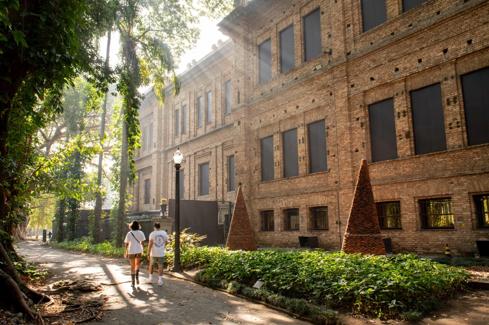 Two people walk on a shaded path beside a large brick building with cone-shaped sculptures in a garden.