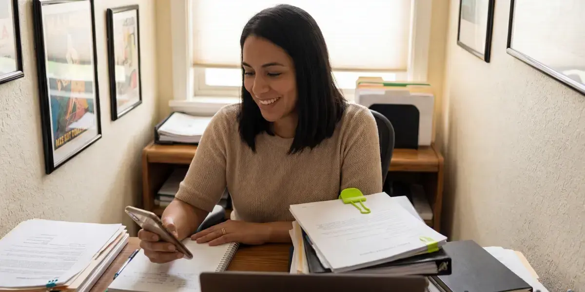 Mulher sorridente analisando no celular quanto rende 100 mil no CDB por mês em seu escritório.